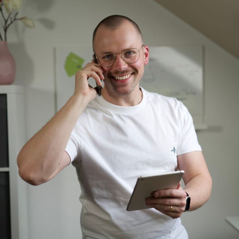 Mann mit kurzen Haaren und einem Lachen und Ipad in der Hand und iPhone am Ohr